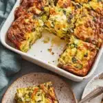 Ultimate Breakfast Casserole with Potatoes 5 Close-up of a slice of savory golden-brown breakfast casserole with potatoes and mixed vegetables, served on a speckled rustic plate.