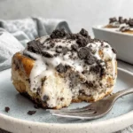 Irresistibly Gooey Oreo Cinnamon Rolls 6 Close-up of a partially eaten, glazed Oreo cinnamon roll topped with white icing and dark cookie crumbles on a light blue speckled plate.