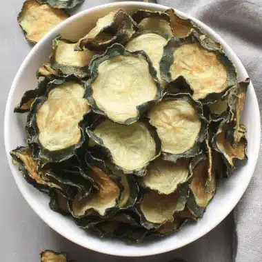 Amazing Crispy Cucumber Chips Recipe 3 Crispy, dehydrated Cucumber Chips in a white ceramic bowl on a grey textured background, highlighting the thin slices and natural green color.