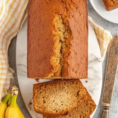 Ultimate One Bowl Banana Bread Recipe 3 Top-down view of a moist, golden-brown loaf of one bowl banana bread on a marble board with two cut slices and ripe bananas.