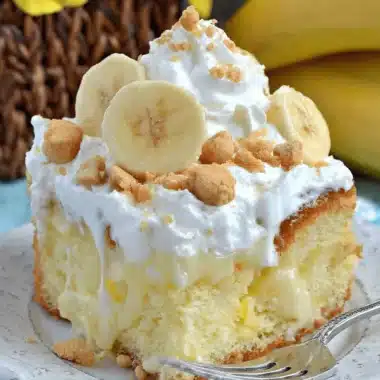 Easy Banana Pudding Poke Cake Recipe 3 A decadent slice of homemade Banana Pudding Poke Cake with whipped cream, fresh banana slices, and crumbled cookies on a rustic plate.