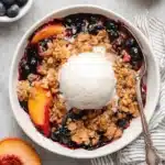 Ultimate Foolproof Blueberry Peach Crumble 7 Delicious Blueberry Peach Crumble with melting vanilla ice cream in a white bowl, surrounded by fresh fruit.