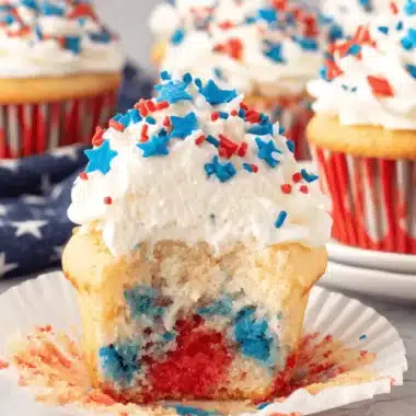 Amazing 4th of July Cupcakes: Easy & Patriotic 4 Close-up of a partially eaten 4th of July cupcake with red and blue marbled cake, white frosting, and patriotic star sprinkles, surrounded by other festive 4th of July cupcakes.