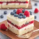 Remarkable American Flag Cake: Stress-Free & Stunning 6 A vibrant slice of American Flag Cake with layers of white cake, red berry filling, and white cream, topped with fresh blueberries and raspberries, served on a wooden plate.