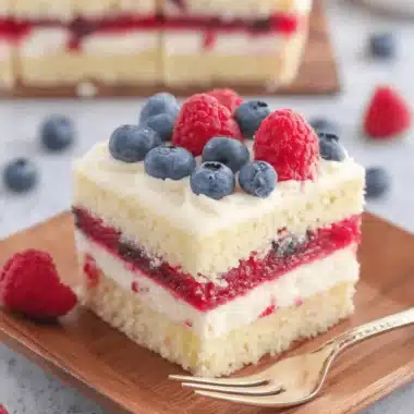 Remarkable American Flag Cake: Stress-Free & Stunning 3 A vibrant slice of American Flag Cake with layers of white cake, red berry filling, and white cream, topped with fresh blueberries and raspberries, served on a wooden plate.