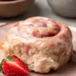 Wonderful, Easy Strawberry Sweet Rolls 6 A single Strawberry Sweet Roll on a pink plate, drizzled with pink glaze, with a halved strawberry beside it and whole strawberries in the background.