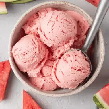 Amazing Watermelon Ice Cream: Your Summer Dream 3 Top-down view of a speckled ceramic bowl filled with scoops of pink watermelon ice cream, an ice cream scoop, and fresh watermelon slices on a light gray surface.