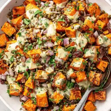 Vibrant Sweet Potato Salad Recipe for Any Meal 4 A vibrant sweet potato salad recipe in a shallow off-white bowl, featuring roasted sweet potatoes, lentils, red onion, dried cranberries, pumpkin seeds, fresh herbs, and a creamy dressing, with silver serving spoons.
