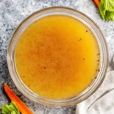 The Ultimate Chicken Stock Recipe 3 A golden bowl of freshly prepared chicken stock, surrounded by bay leaves, carrots, and celery—key ingredients for a delicious chicken stock recipe.