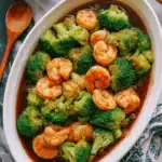 Amazing Honey Garlic Shrimp and Broccoli 5 Close-up of a delicious shrimp and broccoli stir-fry in a white oval dish on a dark surface, ready to eat with chopsticks.
