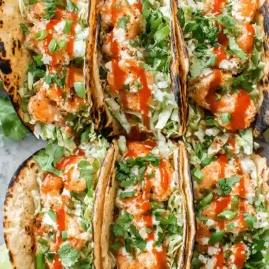 Amazing Bang Bang Shrimp Tacos 4 Six delicious bang bang shrimp tacos, topped with cabbage, seasoned shrimp, creamy sauce, cilantro, and a spicy red drizzle, arranged on a light gray surface.