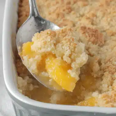Amazing Foolproof Cake Mix Peach Cobbler 3 A silver spoon scoops a generous portion of golden-brown cake mix peach cobbler from a grey baking dish, revealing the juicy peaches underneath.