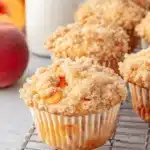 Foolproof Peach Cobbler Muffins: Easy Bakery-Style 8 Close-up of golden-brown peach cobbler muffins with crumble topping on a cooling rack, with fresh peaches and milk in the background.