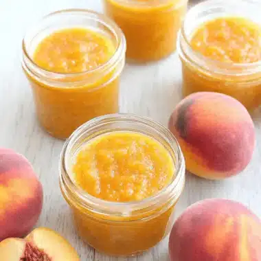 Wonderful Easy Peach Butter Recipe 3 Homemade peach butter in jars with fresh peaches on a wooden surface, highlighting a delicious peach butter recipe.