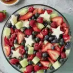 Stunning 4th of July Fruit Salad Recipe 8 Vibrant 4th of July fruit salad in a glass bowl, featuring red strawberries and raspberries, blue blueberries, green melon, and star-shaped fruit, with honey on the side.