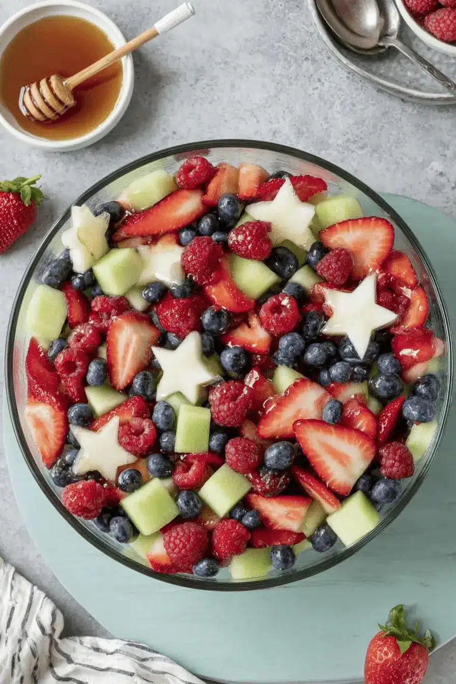 Stunning 4th of July Fruit Salad Recipe 2 Vibrant 4th of July fruit salad in a glass bowl, featuring red strawberries and raspberries, blue blueberries, green melon, and star-shaped fruit, with honey on the side.
