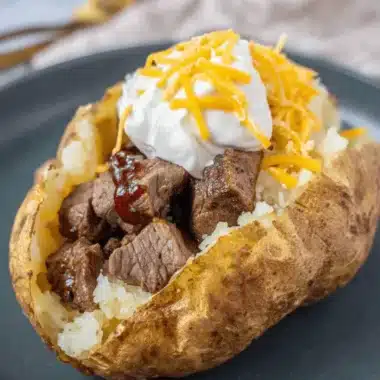The Ultimate Loaded Steak Baked Potato 3 Close-up of a perfectly baked, golden-brown loaded steak baked potato, generously topped with steak tips, sour cream, cheddar cheese, and barbecue sauce.