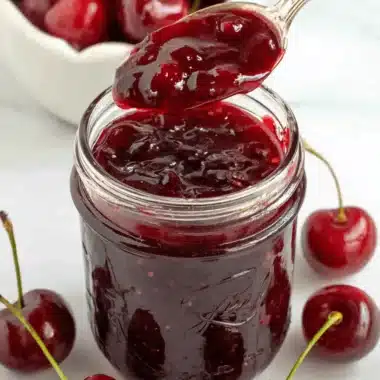 Wonderful Quick batch Cherry Jam Recipe 3 Close-up of a mason jar filled with glistening, deep red batch cherry jam, with a spoon dripping jam back into the jar, surrounded by fresh cherries.