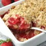 The Ultimate Strawberry Rhubarb Crisp Recipe 8 A close-up of a golden-brown strawberry rhubarb crisp in a white baking dish, with a spoon serving a portion revealing the vibrant fruit filling.
