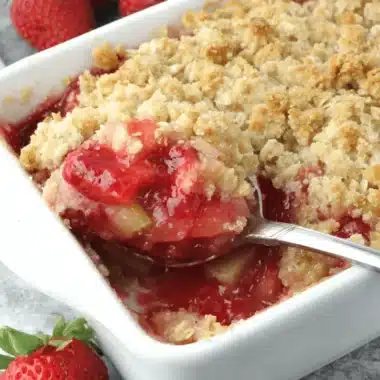 The Ultimate Strawberry Rhubarb Crisp Recipe 4 A close-up of a golden-brown strawberry rhubarb crisp in a white baking dish, with a spoon serving a portion revealing the vibrant fruit filling.