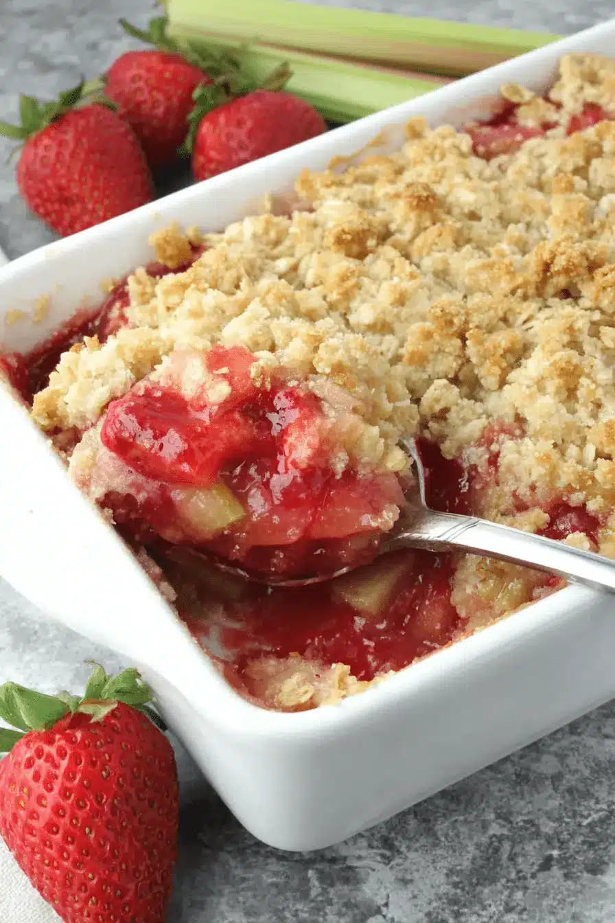 The Ultimate Strawberry Rhubarb Crisp Recipe 2 A close-up of a golden-brown strawberry rhubarb crisp in a white baking dish, with a spoon serving a portion revealing the vibrant fruit filling.