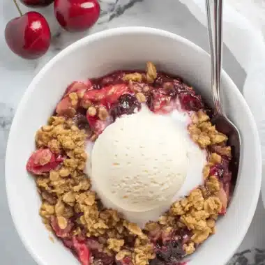 Wonderful Cherry Rhubarb Crisp 4 Top-down view of a delightful cherry rhubarb crisp with a melting scoop of vanilla ice cream in a white bowl, garnished with fresh cherries.