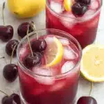 Vibrant Cherry Lemonade: Simple & Refreshing Drink 7 Two refreshing glasses of cherry lemonade with ice, garnished with lemon slices and fresh cherries, on a light surface with scattered whole cherries and lemons.