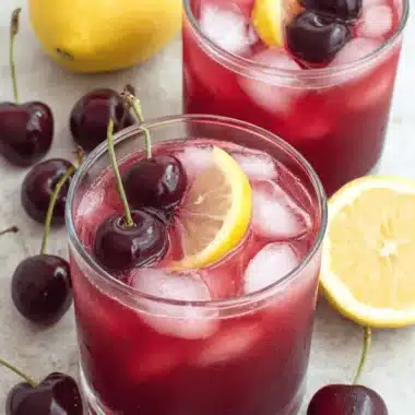 Vibrant Cherry Lemonade: Simple & Refreshing Drink 3 Two refreshing glasses of cherry lemonade with ice, garnished with lemon slices and fresh cherries, on a light surface with scattered whole cherries and lemons.