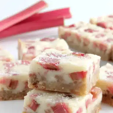 Amazing Foolproof Rhubarb Bars 3 Close-up of freshly baked rhubarb bars with a golden shortbread crust, creamy rhubarb filling, and a dusting of powdered sugar, presented with fresh rhubarb stalks.