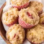 Irresistibly Moist Strawberry Rhubarb Muffins 5 A close-up of freshly baked strawberry rhubarb muffins with a golden-brown streusel topping nestled in a wooden bowl.