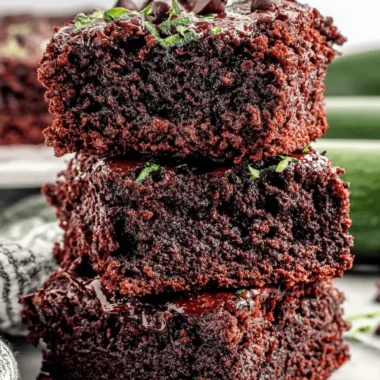 The Ultimate Zucchini Brownies: Fudgy & Foolproof 4 Stack of three rich, glossy dark chocolate zucchini brownies with chocolate chips and subtle green specks on a marble surface.