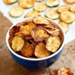 The Ultimate Crispy Baked Zucchini Chips 5 Overhead shot of a deep blue bowl brimming with golden-brown, crispy zucchini chips, with more scattered on parchment paper.
