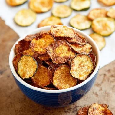 The Ultimate Crispy Baked Zucchini Chips 4 Overhead shot of a deep blue bowl brimming with golden-brown, crispy zucchini chips, with more scattered on parchment paper.