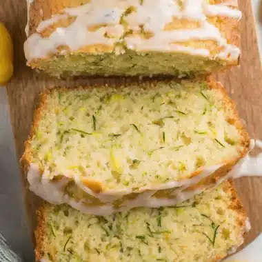 Ultimate Foolproof Lemon Zucchini Bread 3 Slices of moist lemon zucchini bread with white glaze and a fresh lemon slice on a wooden board.