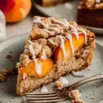 Amazing Peach Coffee Cake: Easy & Moist 7 A close-up of a delicious slice of peach coffee cake with a crumble topping and white glaze on a speckled plate.