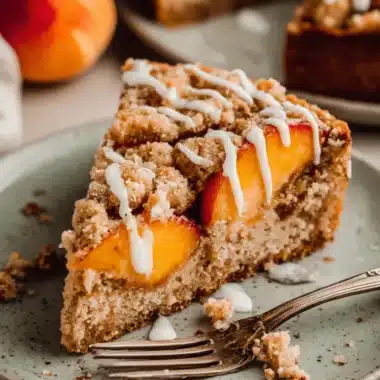 Amazing Peach Coffee Cake: Easy & Moist 3 A close-up of a delicious slice of peach coffee cake with a crumble topping and white glaze on a speckled plate.