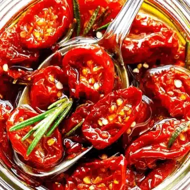 Fantastic Oven Sun Dried Tomatoes: Easy Homemade 3 A close-up, top-down view of a glass jar filled with vibrant oven sun dried tomatoes, preserved in golden oil with fresh rosemary, a spoon ready to serve.