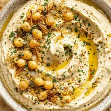 Amazing Homemade Hummus: Easy, Creamy Perfection 3 Creamy homemade hummus garnished with chickpeas, caramelized onions, chives, red pepper flakes, and olive oil, served with pita bread.