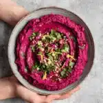 Amazing Vibrant Beet Hummus Recipe 7 Vibrant beet hummus in a rustic bowl, garnished with fresh herbs and olive oil, held by hands.