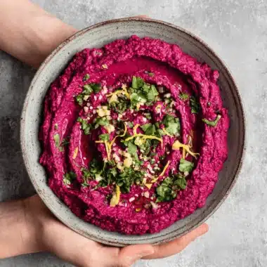 Amazing Vibrant Beet Hummus Recipe 4 Vibrant beet hummus in a rustic bowl, garnished with fresh herbs and olive oil, held by hands.