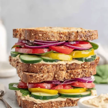 Amazing Hummus Sandwich: Quick & Easy Lunch 3 A tall, multi-layered whole-grain hummus sandwich with red onion, tomatoes, cucumbers, and spinach, served on a light gray surface with a bowl of hummus.