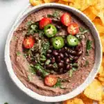 Amazing Creamy Black Bean Hummus Recipe 6 Top-down view of homemade black bean hummus garnished with cherry tomatoes, jalapeños, cilantro, and served with tortilla chips and red bell peppers.