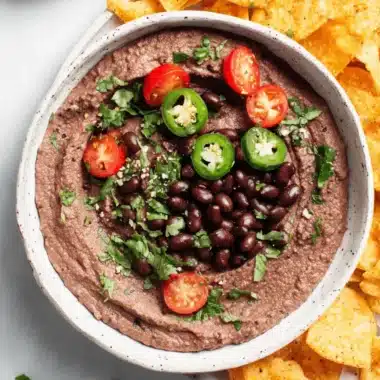 Amazing Creamy Black Bean Hummus Recipe 4 Top-down view of homemade black bean hummus garnished with cherry tomatoes, jalapeños, cilantro, and served with tortilla chips and red bell peppers.