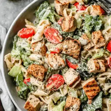 The Ultimate Ceasar Pasta Chicken Salad 3 A close-up, overhead view of a hearty ceasar pasta chicken salad with grilled chicken, cherry tomatoes, and romaine lettuce in a light gray bowl.