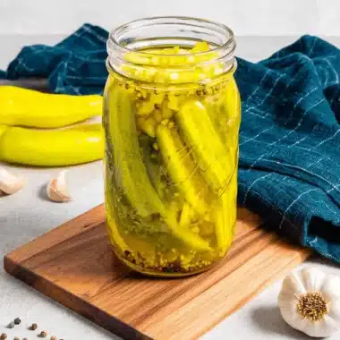 Amazing Pickled Banana Peppers: Easy Homemade Recipe 3 A mason jar of bright yellow pickled banana peppers with spices on a cutting board, alongside fresh peppers and garlic.