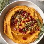 Amazing Easy Pumpkin Hummus Recipe 5 Vibrant orange pumpkin hummus swirled and topped with bright pomegranate seeds, green pepitas, and a sprig of fresh rosemary.
