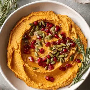 Amazing Easy Pumpkin Hummus Recipe 4 Vibrant orange pumpkin hummus swirled and topped with bright pomegranate seeds, green pepitas, and a sprig of fresh rosemary.