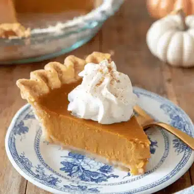 The Ultimate Foolproof Pumpkin Pie Recipe 3 A beautifully presented slice of homemade pumpkin pie recipe, topped with whipped cream and cinnamon, on a patterned plate with a golden fork.