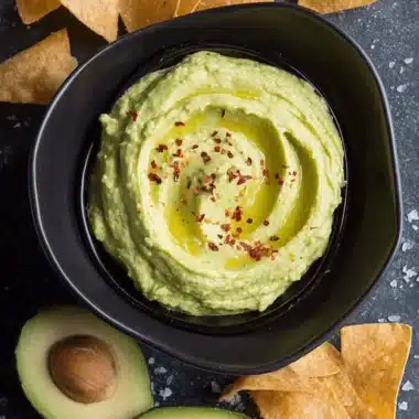 Unbelievably Creamy Avocado Hummus Recipe 3 Creamy avocado hummus garnished with olive oil, red pepper flakes, fresh avocado, and tortilla chips on a dark stone surface.