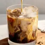 Amazing Homemade Iced Pumpkin Chai Tea Latte 5 A refreshing iced pumpkin chai tea latte being prepared, with thick cream pouring into a glass filled with ice and the dark tea mixture, creating beautiful swirls on a wooden surface with spices.